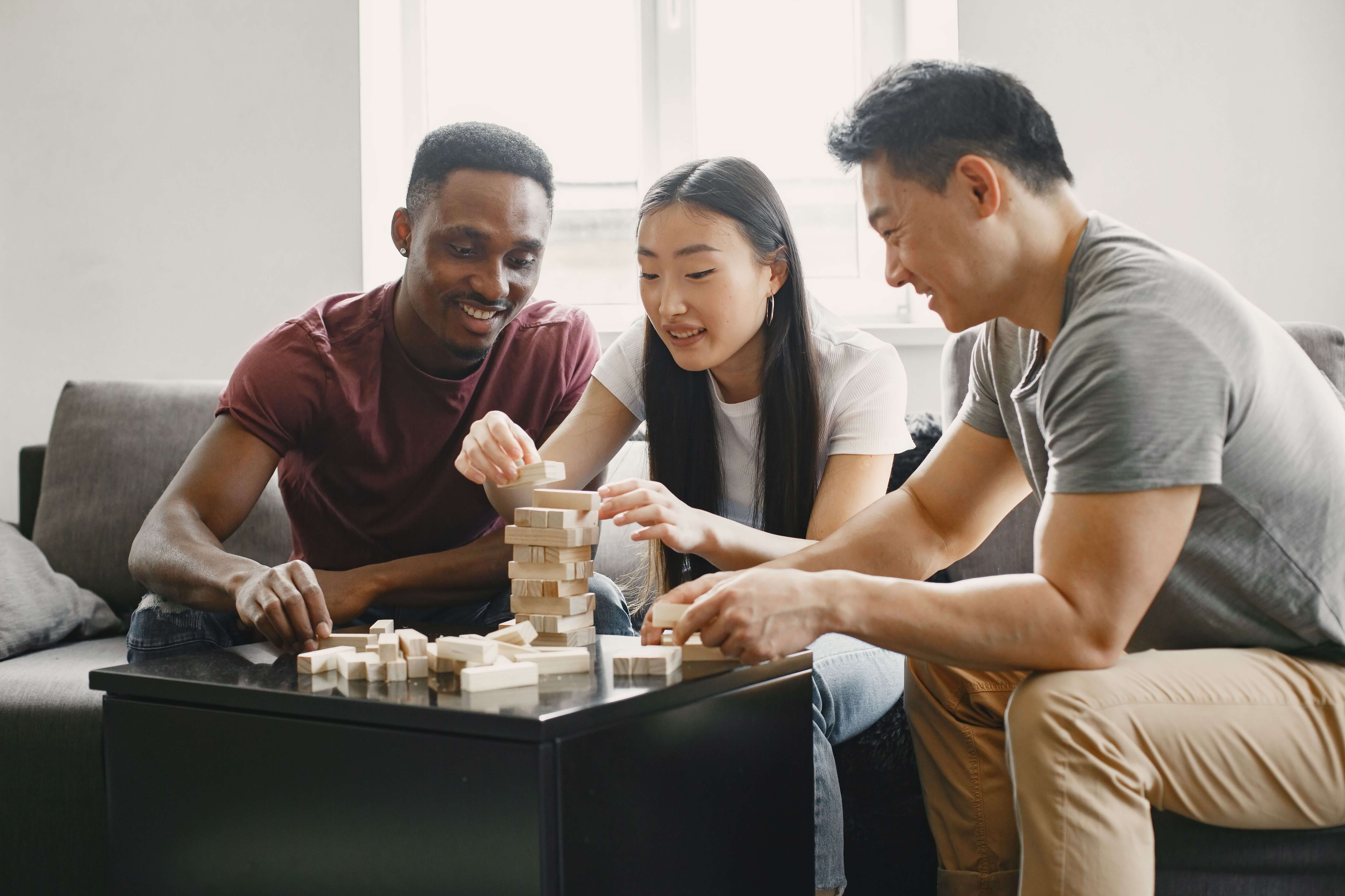 Three friends of Black and East Asian heritage laughing over a game of stacking blocks
