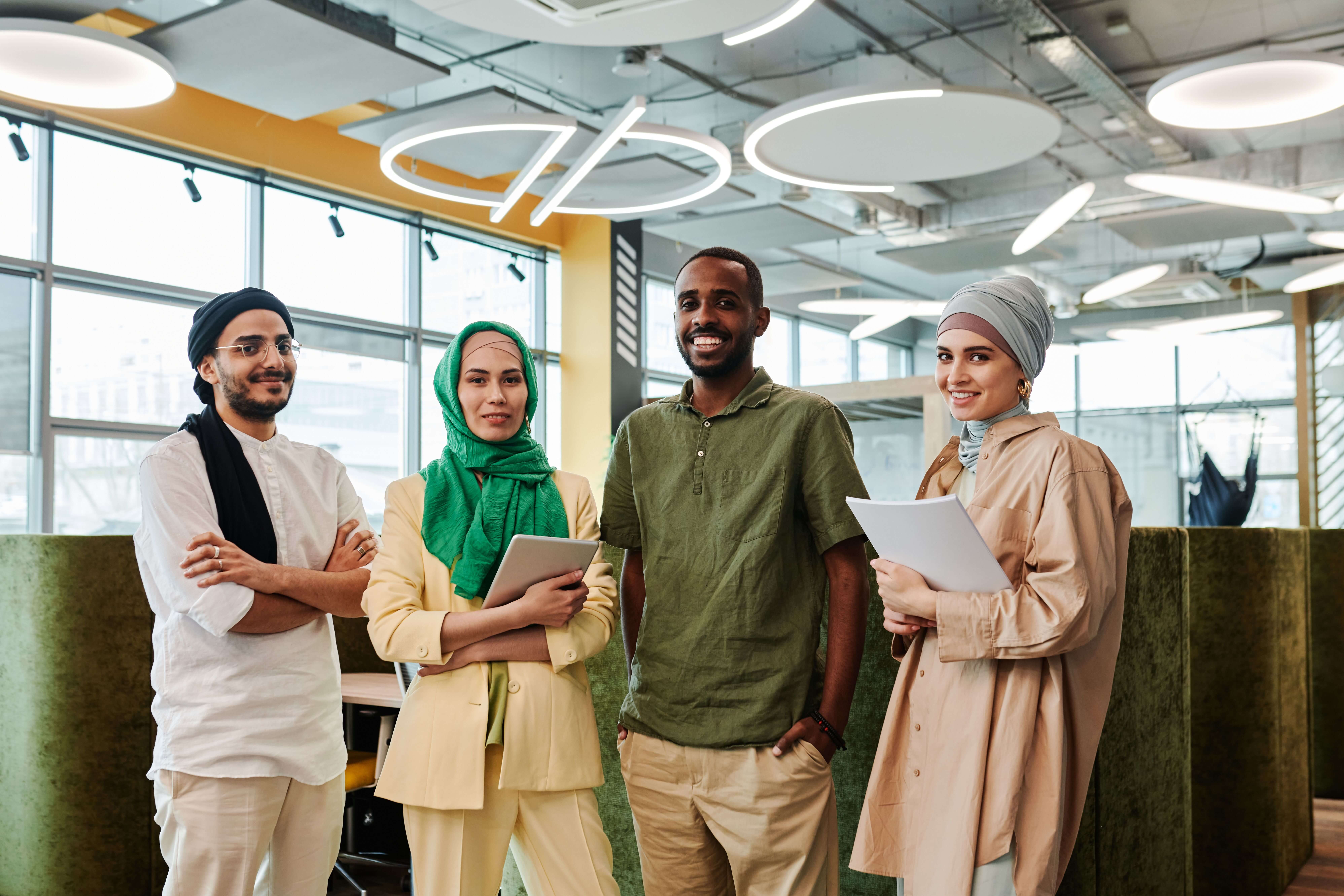Four smiling young colleagues from different backgrounds standing together in a bright office, holding a tablet and papers